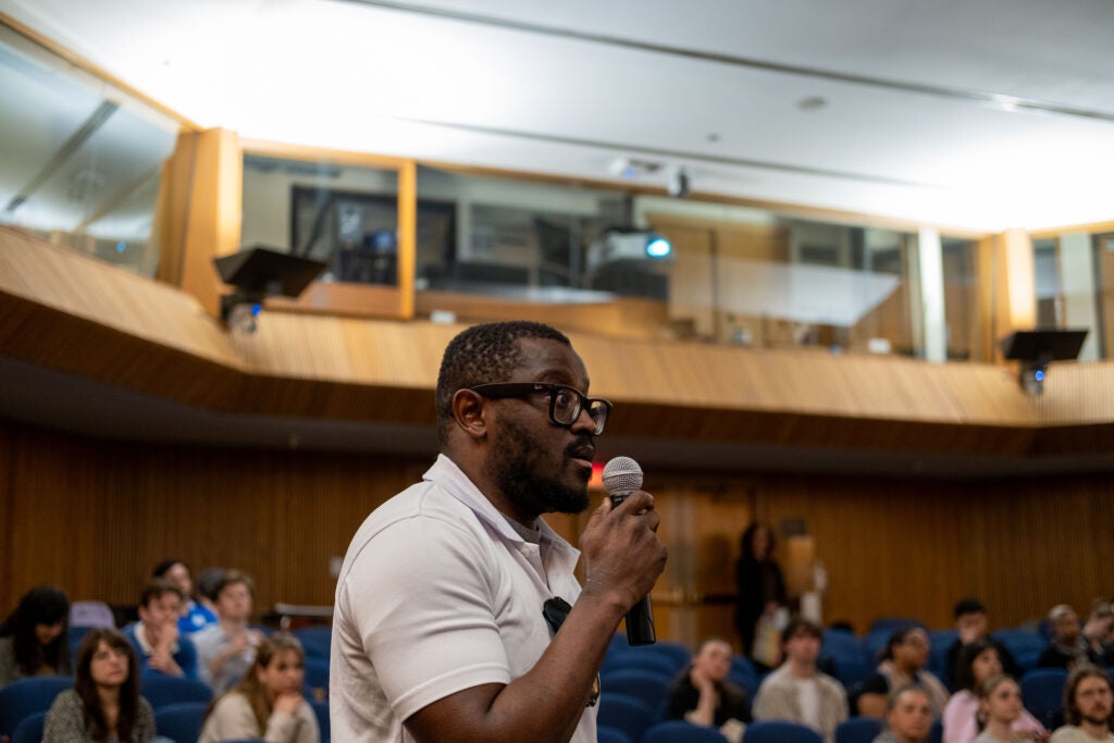 The side profile of an audience member standing and speaking into a microphone. He is wearing glasses and a short sleeve white polo shirt.