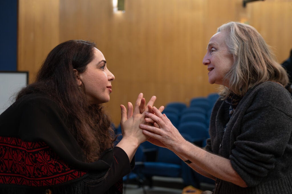 The side profile of two women talking. They each have their hands clasped in front of them, barely touching each other’s hands. The woman on the right is speaking, and the woman on the left looks closely at her. Behind them, the audience seats are empty.