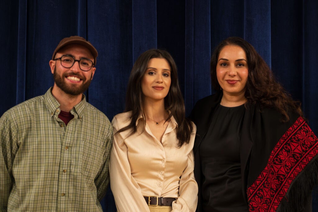 The three panelists pose and smile for a photo. On the left is Layth Malhis, wearing a green plaid button down shirt, black square glasses, and a brown baseball cap. In the middle is Sarah Ramourg, wearing a silk cream-color button down shirt with her hands clasped in front of her. On the right is Tamara Taha, wearing a black shawl with red Palestinian style embroidery along the edge.
