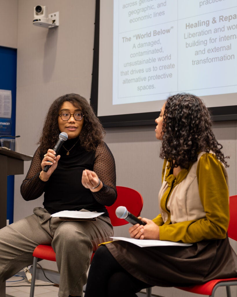Two women sitting in red chairs in front of a projector screen. They are both holding microphones. The woman on the left with square rimmed glasses is speaking and holds one hand in front of her, while the woman on the right looks towards her and holds the microphone in her lap.

