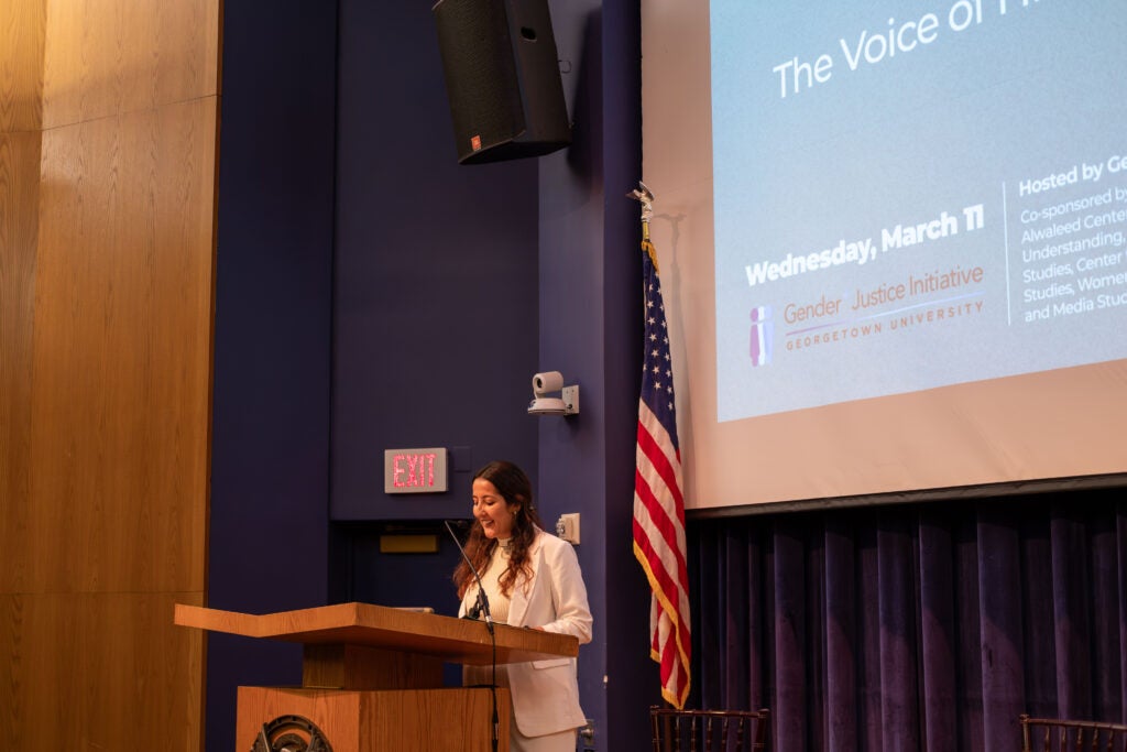A woman wearing a white blazer stands behind a podium, speaking into a microphone. Behind them is an American flag and a projector screen.