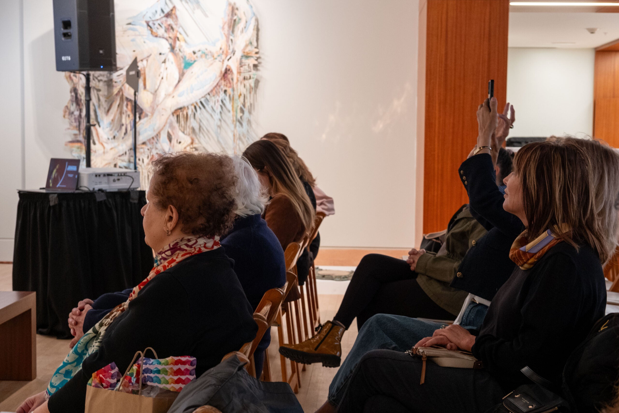 The side profile of a seated audience facing the left. One audience member raises their cellphone in the air and takes a photo. A table in the back left holds a projector screen and a laptop computer.