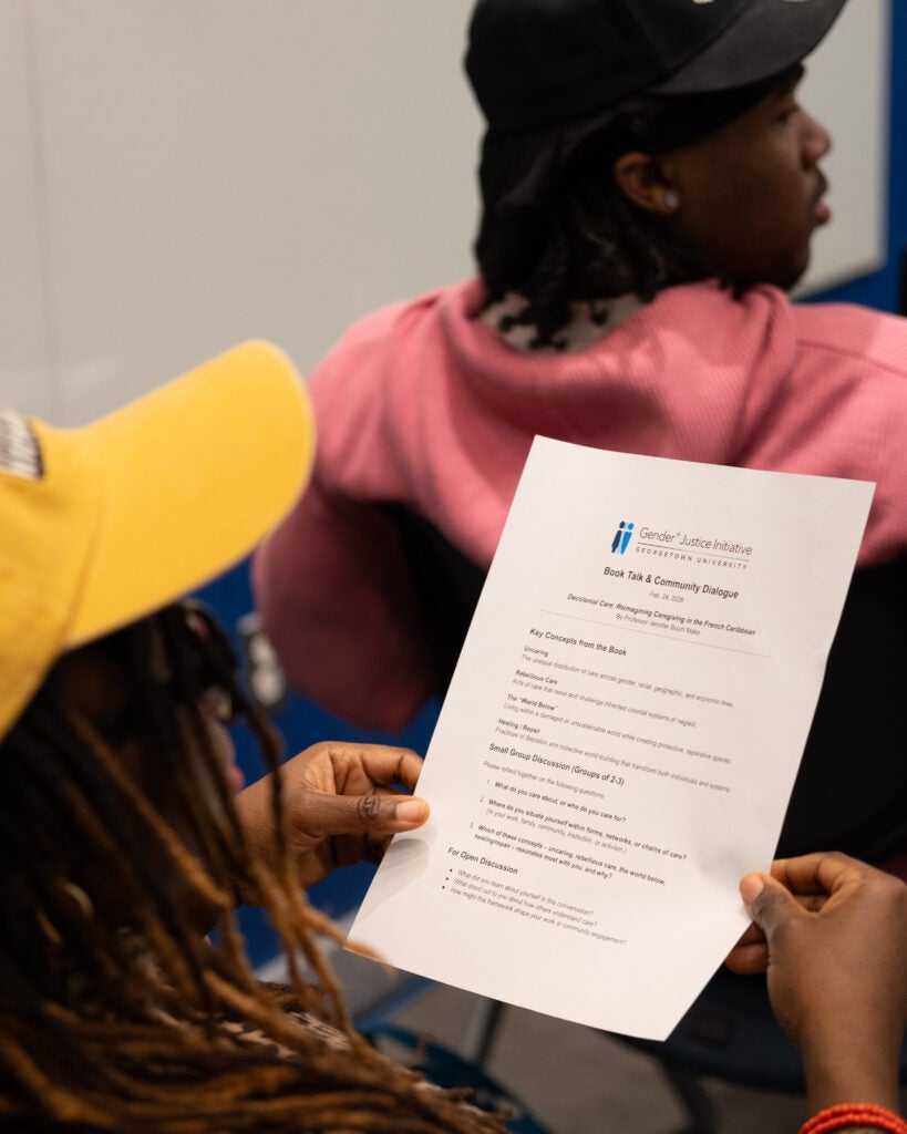 An overhead shot of a woman in a yellow baseball cap holding a piece of paper with both hands. The paper features the Gender+ Justice Initiative logo at the top and the title reads “Book Talk & Community Dialogue” with a list of discussion questions.