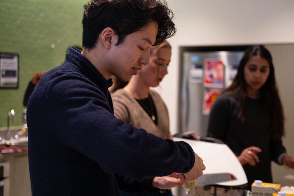 A man opening a can of tomato paste. Behind him, one woman slides items off a cutting board with a knife, and another woman watches.