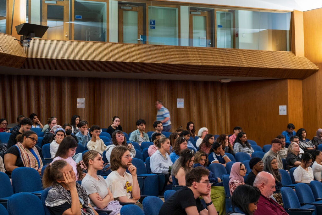 A wide angle photo of the seated audience from the side, looking towards the stage. Some have their head held in their hands. 