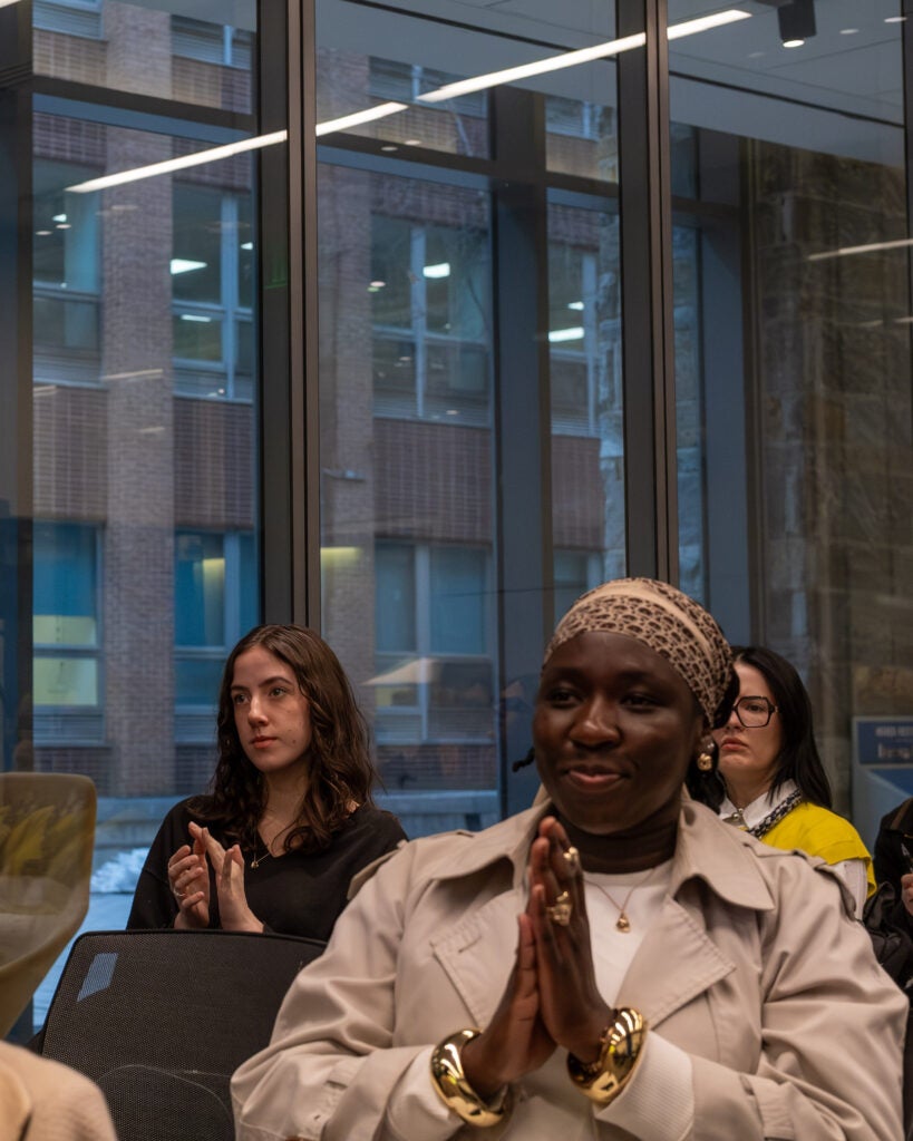 Three seated audience members facing forward and applauding. The woman in the front is wearing gold bangle bracelets, rings, and earrings. 