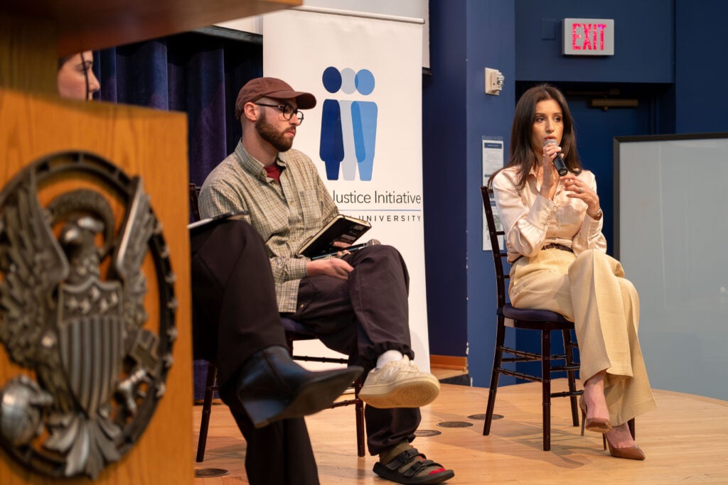 Close up photo of two women and one man sitting on a stage. The woman on the right is speaking into a microphone. Behind her is a banner displaying the Gender+ Justice Initiative logo. A podium on the far left covers up the woman on the left except her face.