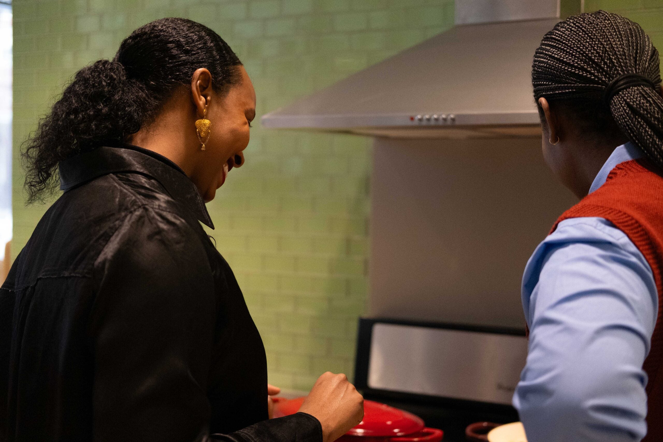 The back of two women standing over a red pot on the stovetop. The woman on the left is smiling. 