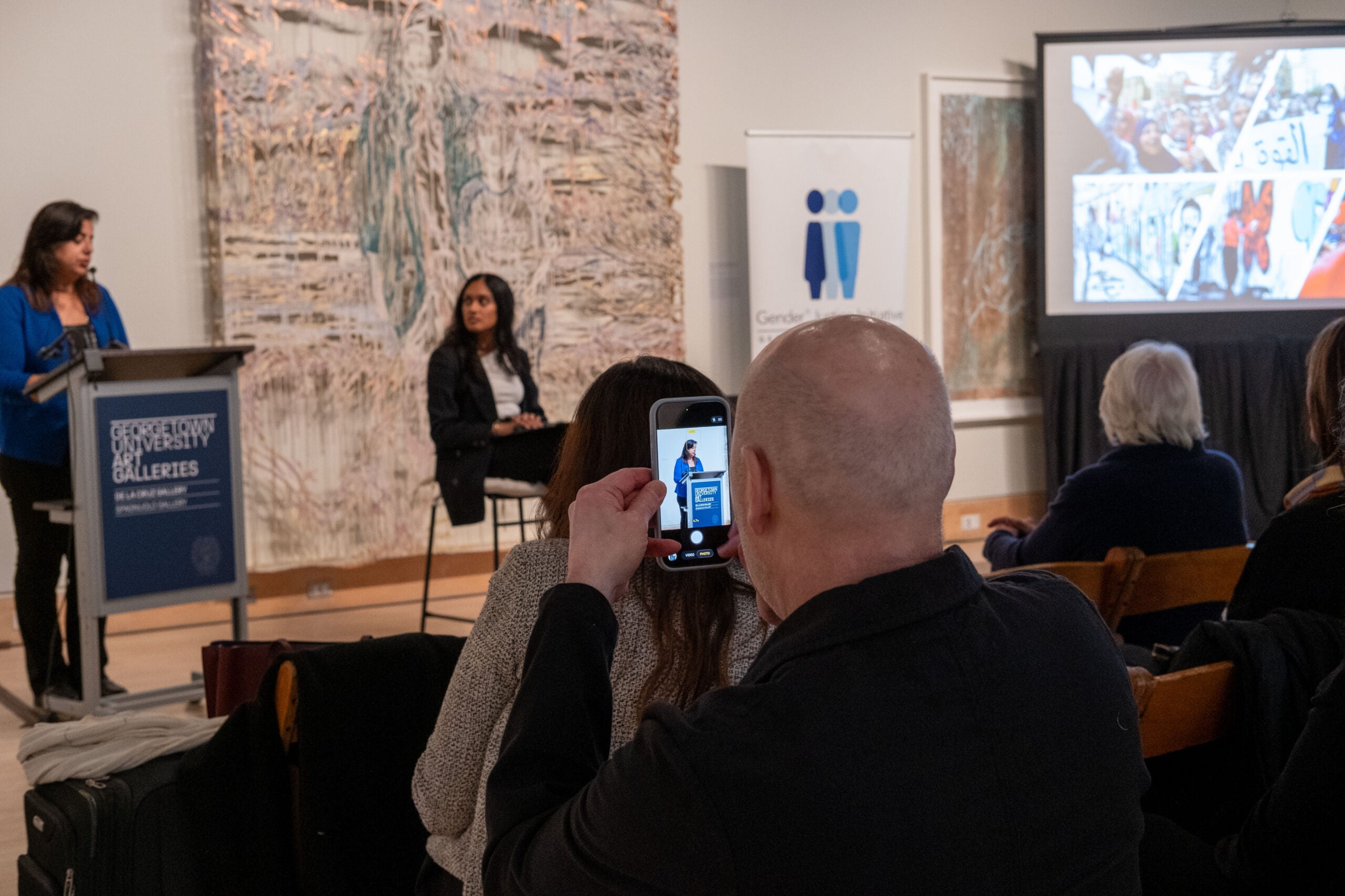 A woman stands behind a podium talking, and another woman is seated to the left, looking at her. A seated audience watches them. A man in the audience holds up a cellphone and takes a picture of the woman at the podium.