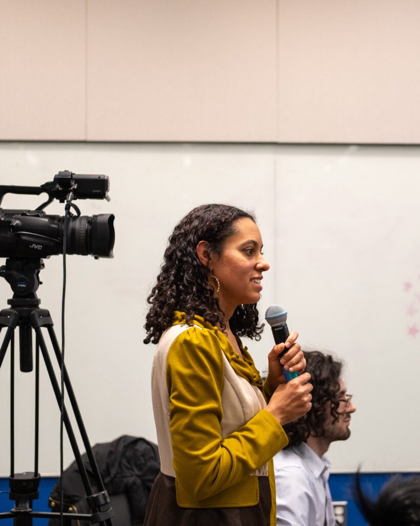 The side profile woman in a long sleeve yellow and beige shirt looking right, standing, and holding a microphone. On the left behind her is a video camera on top of a tripod.