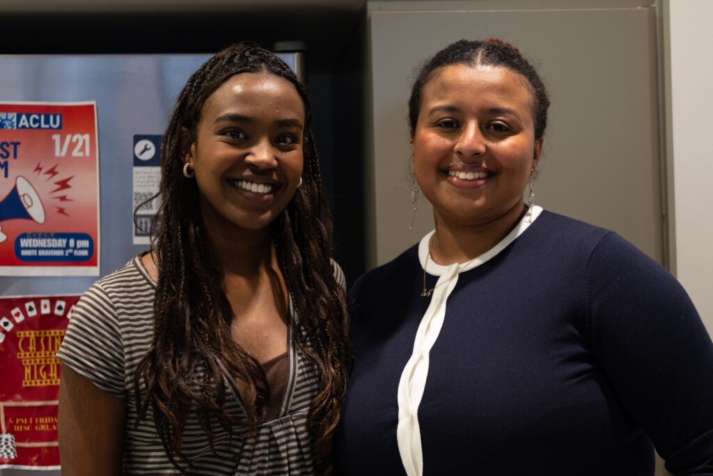 Two women smiling for a photo. Behind them is a stainless steel refrigerator with several colorful event posters taped to it.
