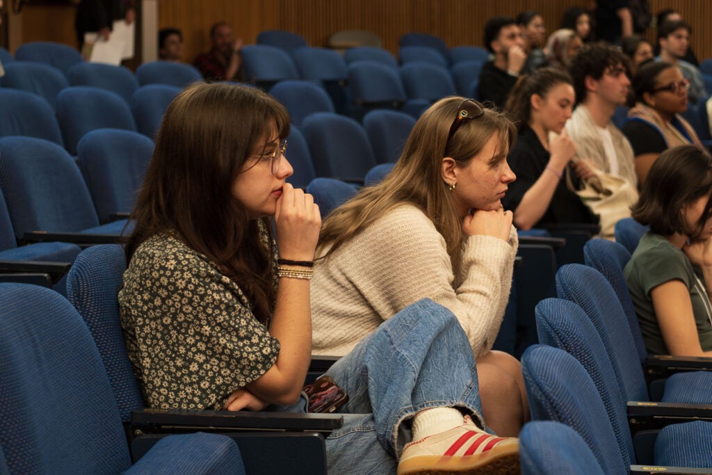 Two women in the audience. The woman on the left has her legs crossed and holds a hand to her chin. The woman on the right is leaned forward, elbows on her knees, head propped up on her hands.
