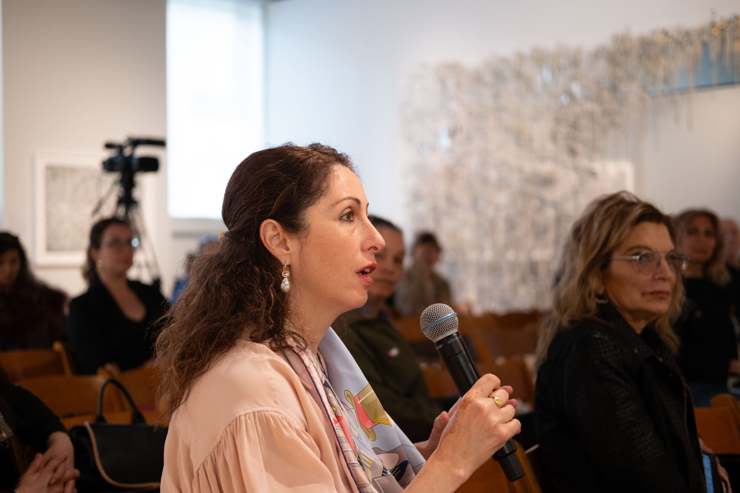 A woman speaking into a microphone, holding it with both hands. Some members of the audience look at her, and others look forward. A camcorder is sitting on a tall tripod in the back of the room, facing forward.