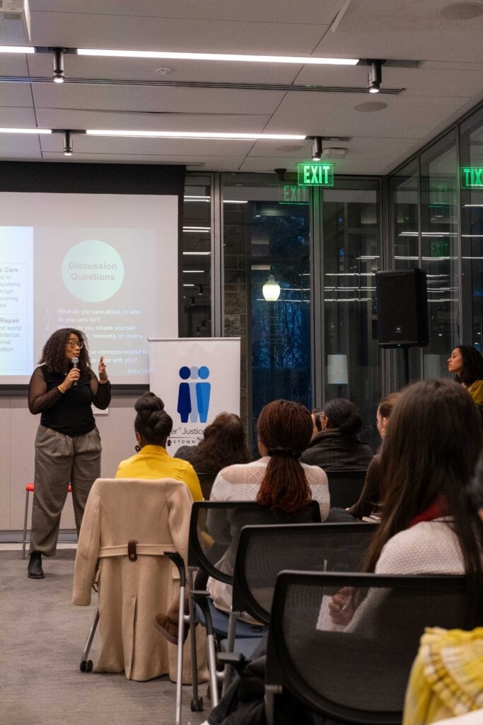 A photo from the back of a room, showing a seated audience facing forward. In front of them is a woman with large black rimmed glasses standing and speaking into a microphone. To the right of her is a banner displaying the Gender+ Justice Initiative logo, and behind her is a large projection screen displaying a slideshow presentation.