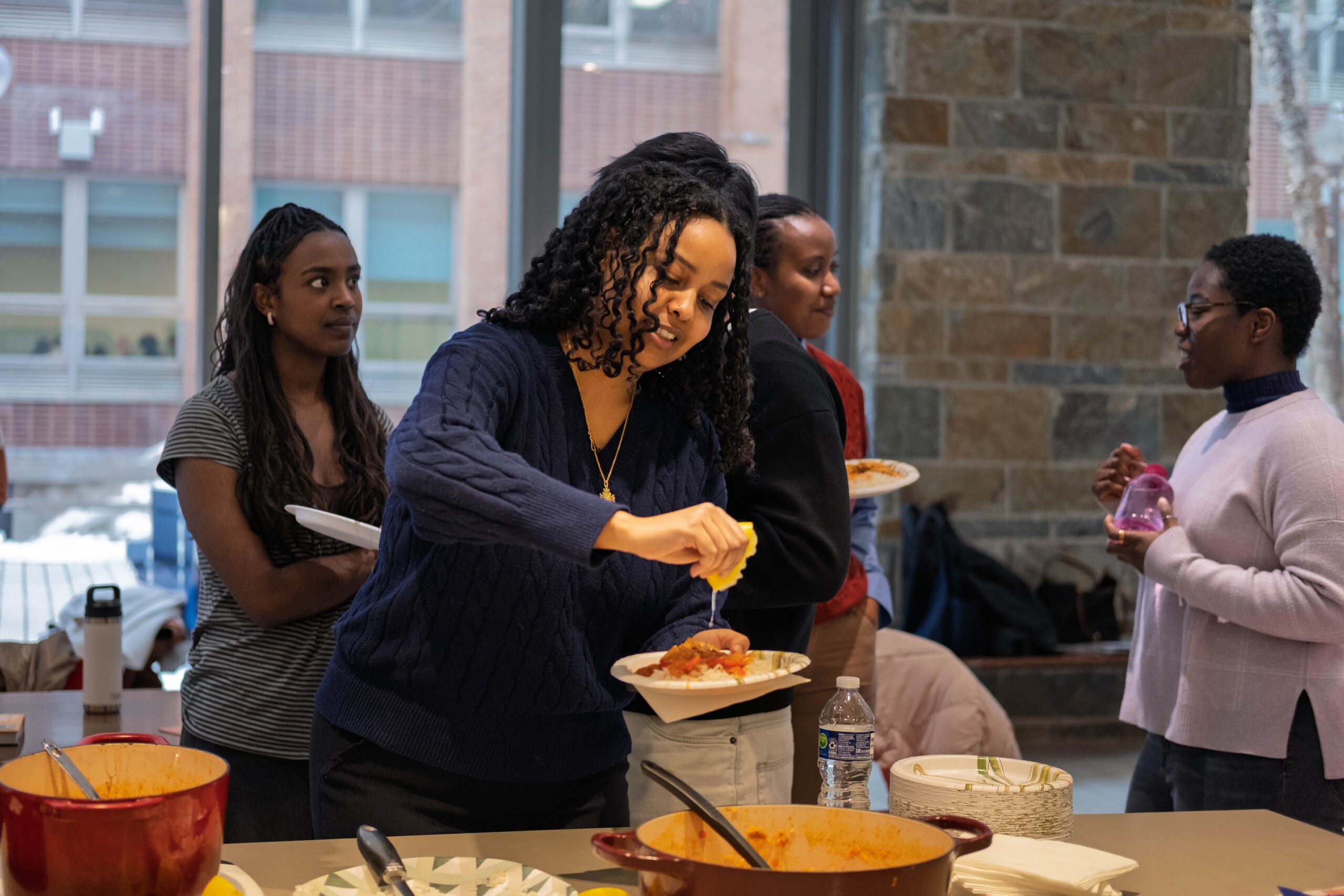 A woman squeezing lemon juice onto a plate of food. Behind her in line, three other women hold plates and converse. 