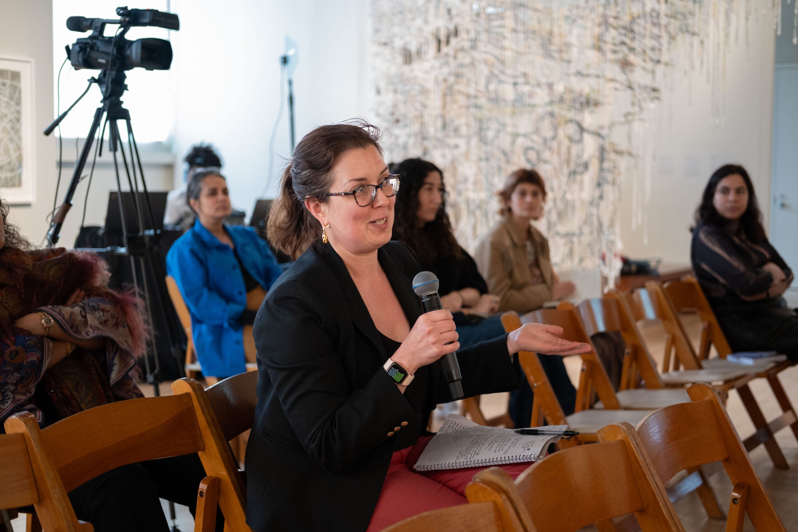 A woman speaking into a microphone, holding it with one hand and gesturing in front of her with the other hand. She is smiling and has a notebook full of writing resting on her lap.