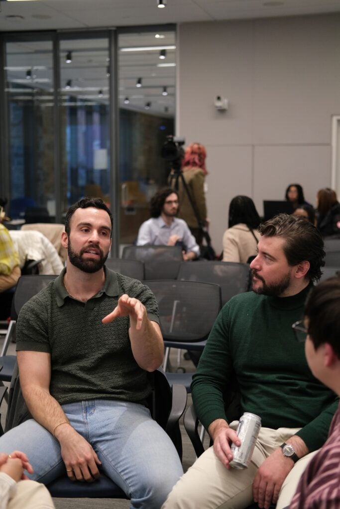  A group of audience members discussing while sitting in a circle. The man on the left is speaking and holding up a hand in front of him, palm-down. The man on the right looks towards him and holds a canned drink in his hand.