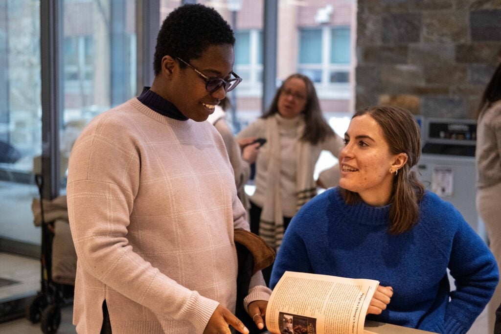 A woman standing up and flipping through a brochure on the counter in front of her. To her right, another woman sits and smiles, in mid-conversation with her.