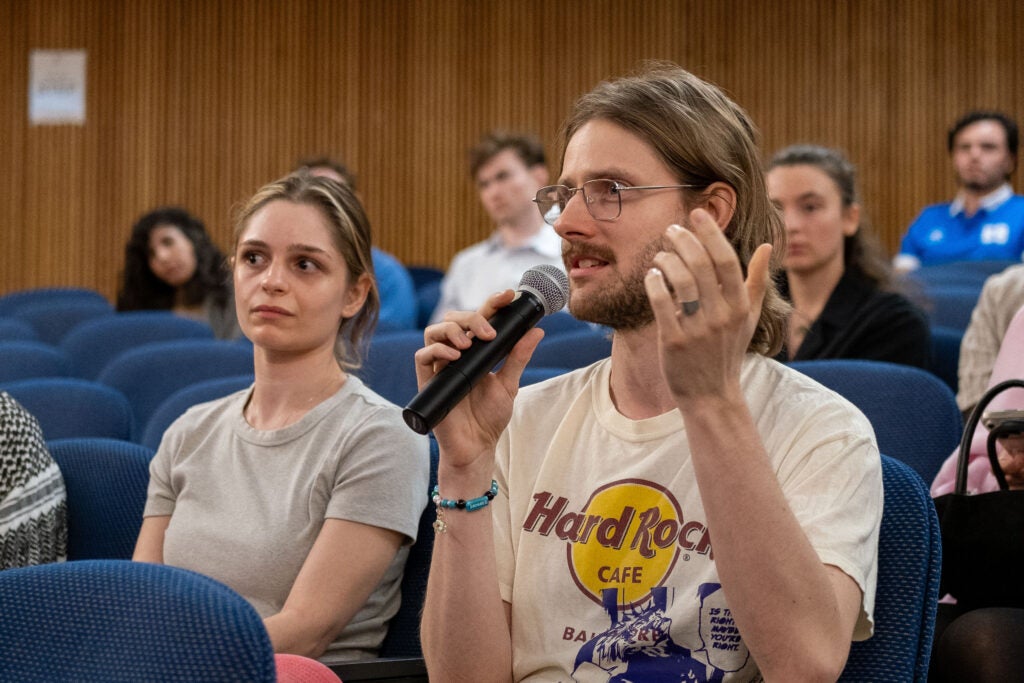 A seated audience member speaks into a microphone. He is gesturing into the air with his other hand. The woman next to him looks intently towards the stage. Audience members in the background are looking towards the man with the microphone.