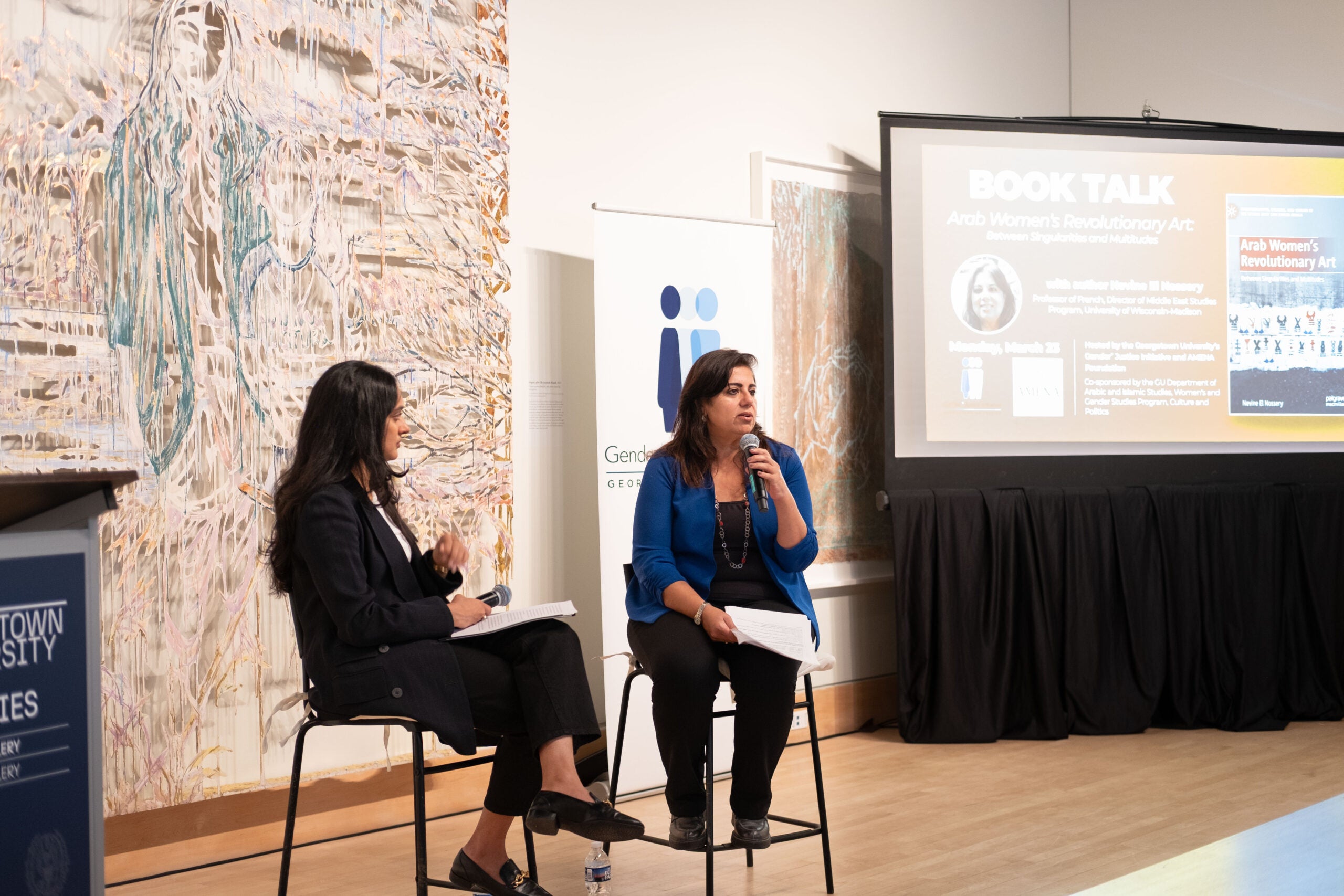 Two women sitting on stage. The woman on the right is speaking into a microphone and holding several sheets of paper. Behind her is the Gender+ Justice Initiative banner and a projector advertising the event.