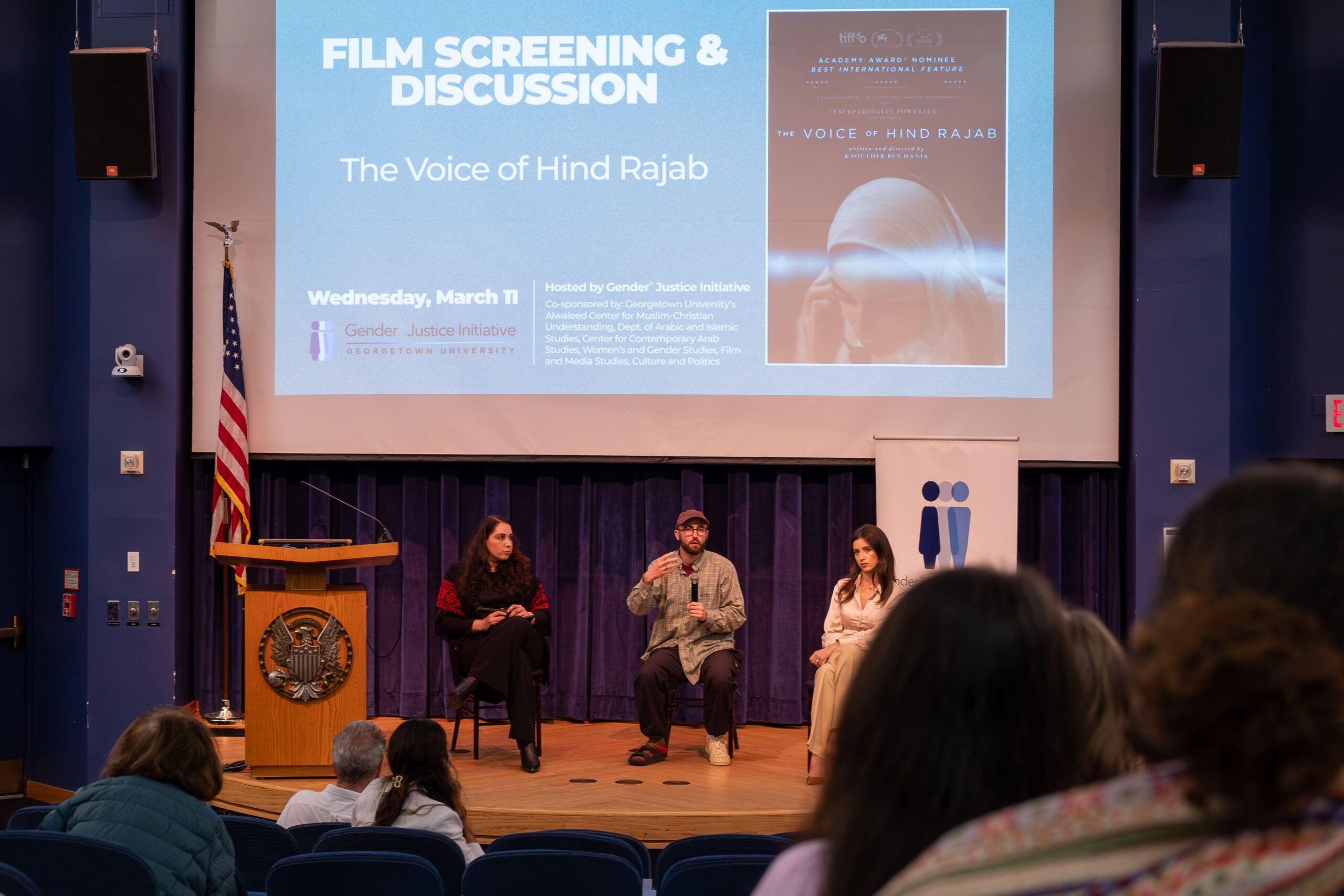 Three people sitting on a stage. The man in the middle is speaking into a microphone. Behind them a large projector screen displays the poster for the film The Voice of Hind Rajab.