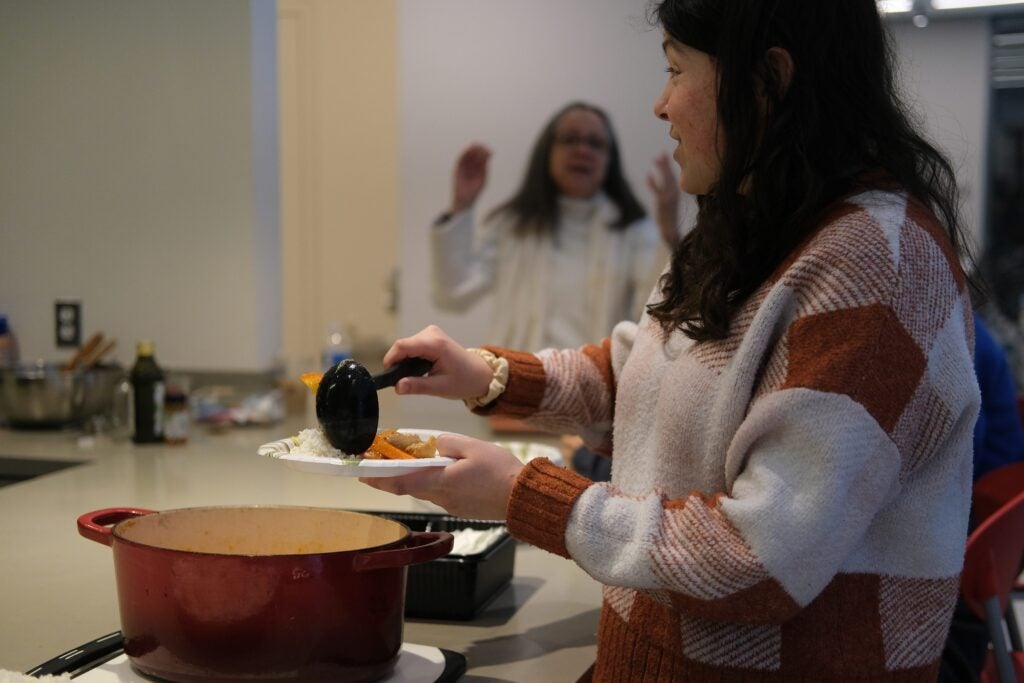 The side profile of a woman scooping stew from a large red pot onto a paper plate using a ladle.