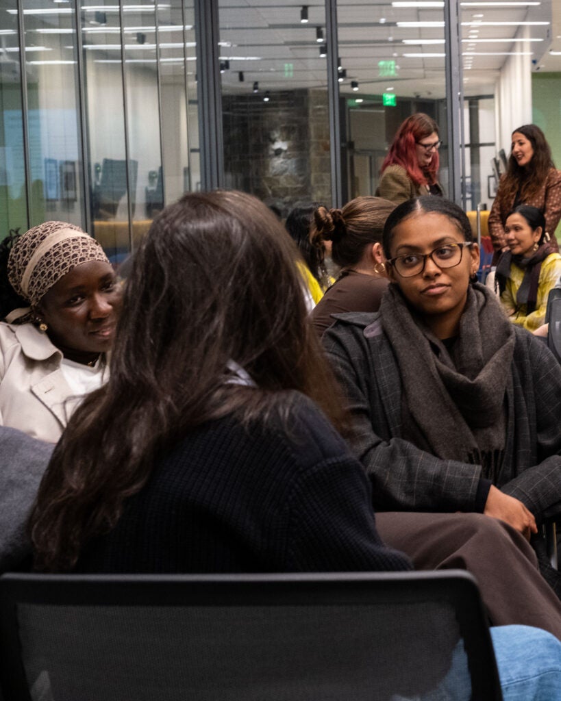A group of audience members discussing while sitting in a circle. The woman in the middle has her back turned to the camera, speaking to two women facing her. Other audience members are sitting in small groups behind them.