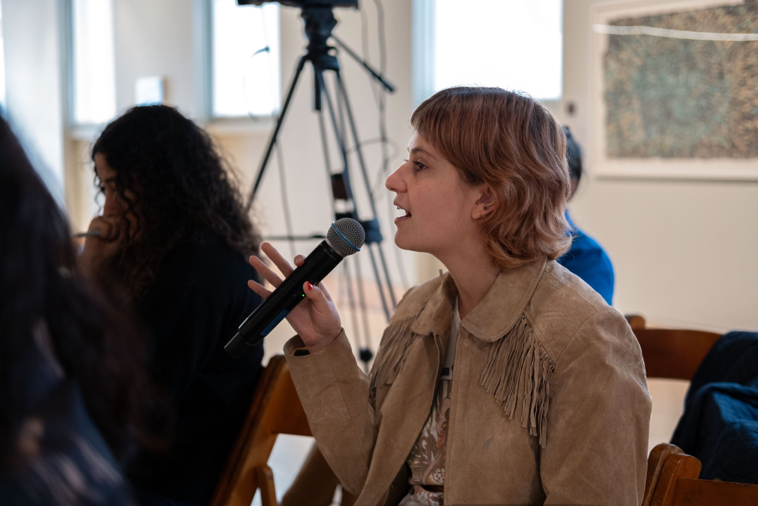 A woman speaking into a microphone, holding it between her thumb and index finger, with her other three fingers pointed outwards. The woman next to her holds a pen in her fist up to her mouth and looks downwards.