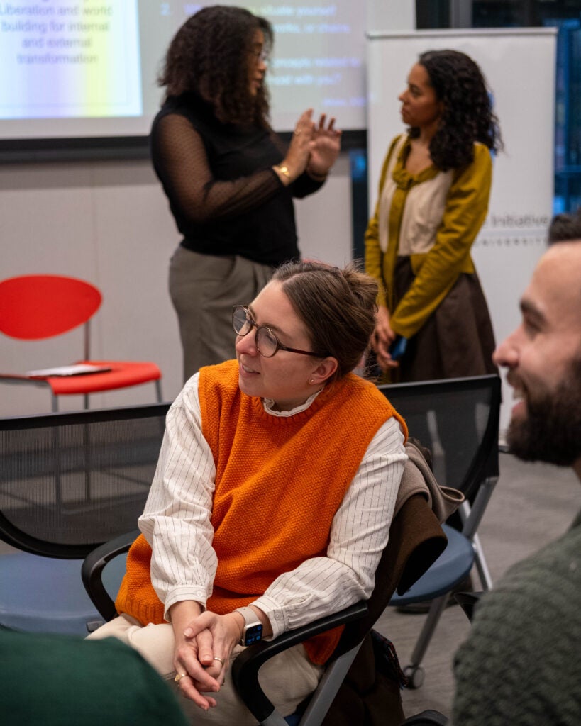 A group of audience members discussing while sitting in a circle. The woman in the middle is wearing a bright orange sweater vest. She has her hands clasped in her lap and is looking to the left with a smile on her face. Behind her, two women are standing in front of a projector screen and talking.