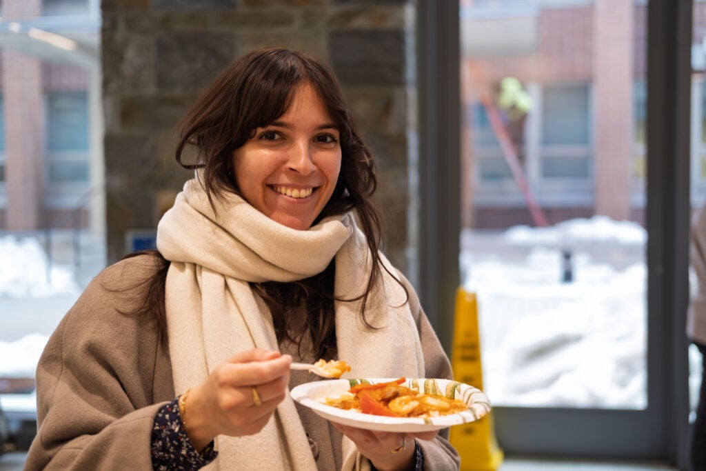 A woman wearing a large white scarf smiling and holding a spoonful of food in one hand, and her plate in the other.
