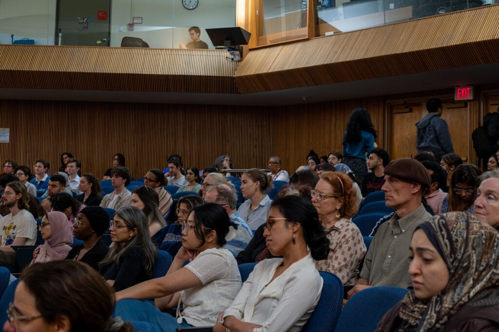 A wide angle photo of the seated audience from the side, looking towards the stage. Two people are walking down the center aisle to the back of the room.