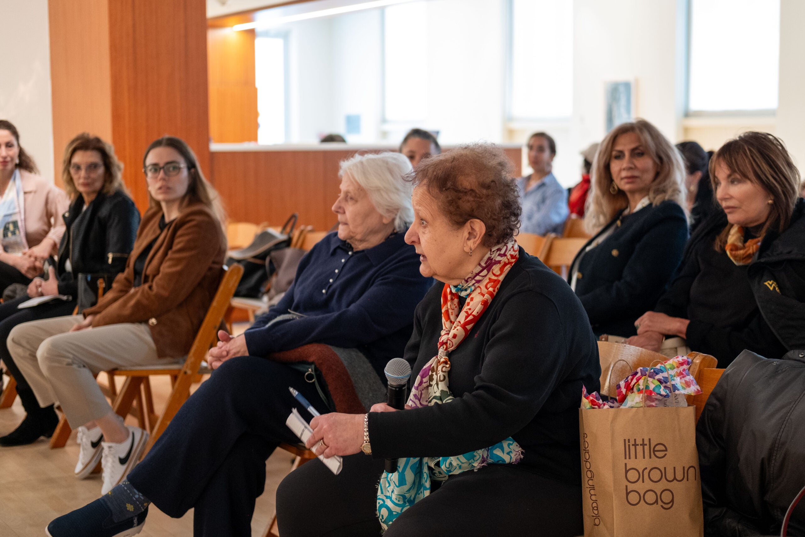 A woman in the front row of the audience speaking and holding a microphone in her lap. The first two rows of audience members look towards her as she speaks.