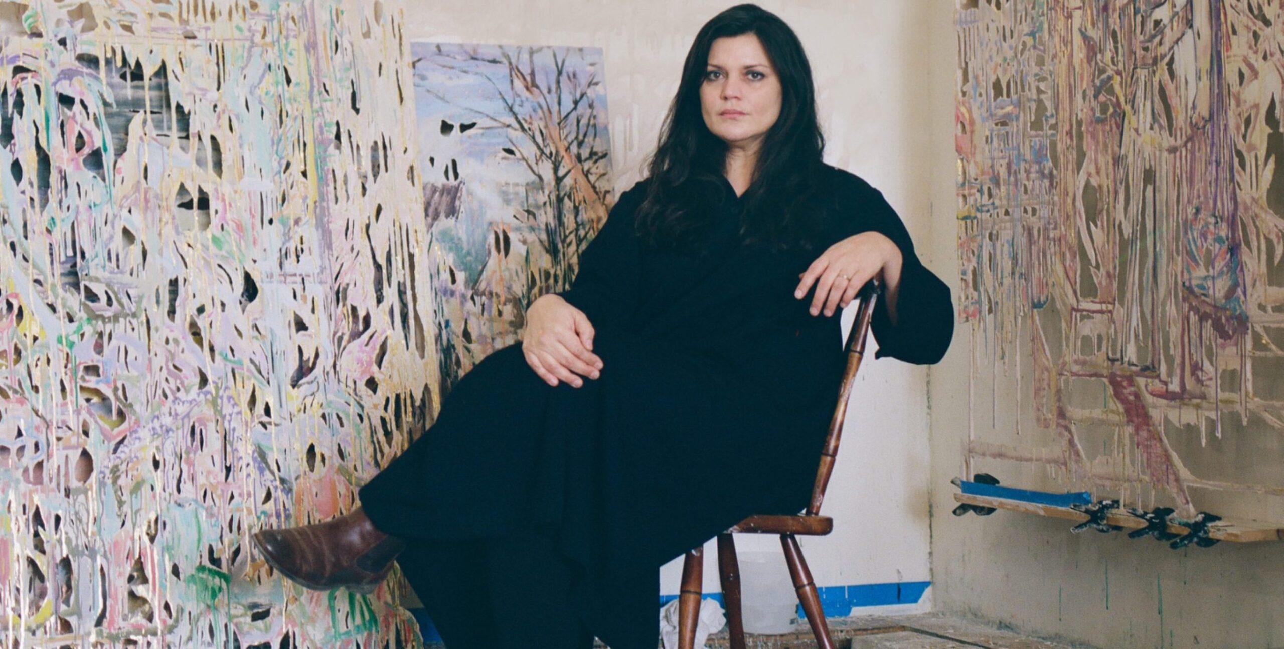 Portrait of artist Diana Al-Hadid seated on a wooden chair in her studio, wearing a long black dress and brown boots. She poses calmly amid large, textured abstract artworks leaning against the walls around her.