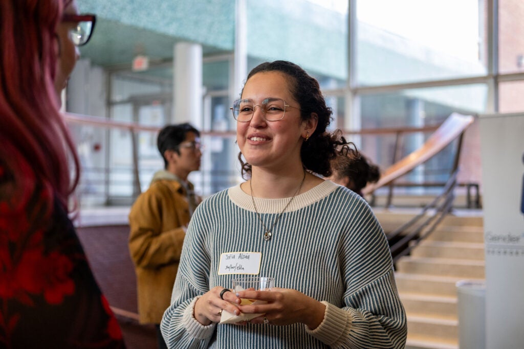 A woman with a nametag sticker on their shirt talking to someone standing to the left. She is smiling and holding a cup in front of her. Behind her, others talk in groups.