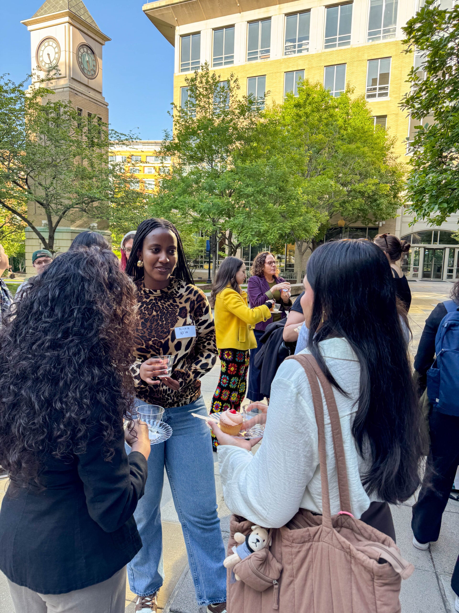Three women standing outside in a circle, talking and holding cups and plates. Others stand and chat behind them.