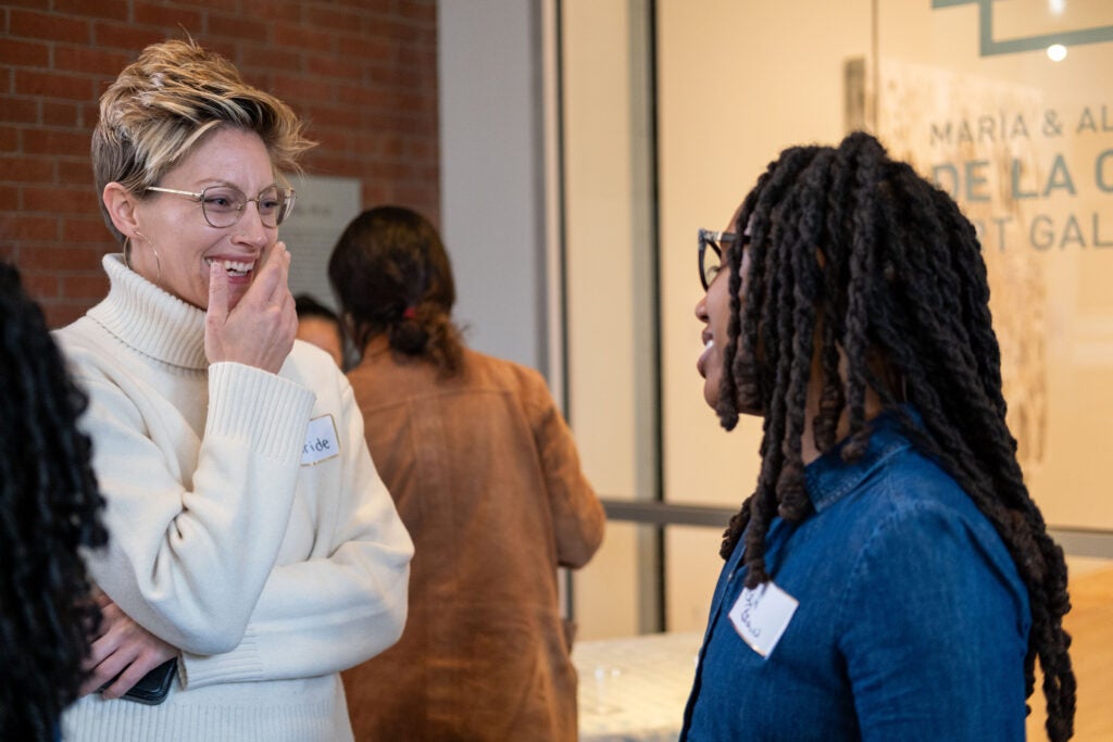 Two women talking to each other. The woman on the left is laughing and holding a hand to her mouth, with her other arm crossed in front of her, holding her phone. The woman on the right is smiling with her eyes closed.