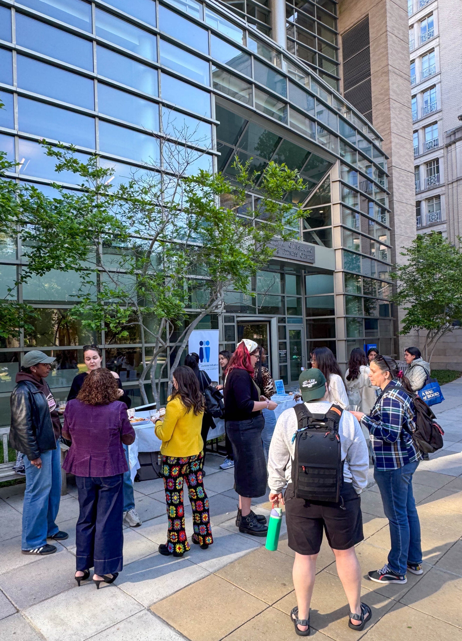 Several people standing outside, talking to each other and holding snacks and cups in their hands. Behind them is a large, tall building with a wall of windows.