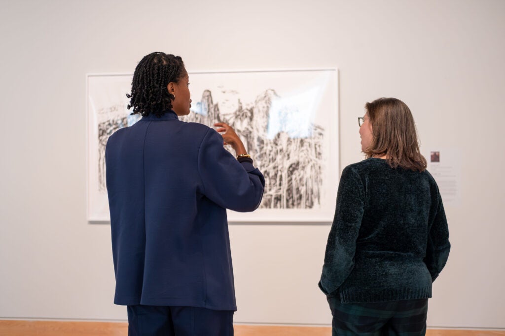 The back of two women looking at a piece of art hung on a museum wall. The woman on the left has her hands gestured up in the air and is talking. The woman on the right has her hands in her pocket and looks towards the art.