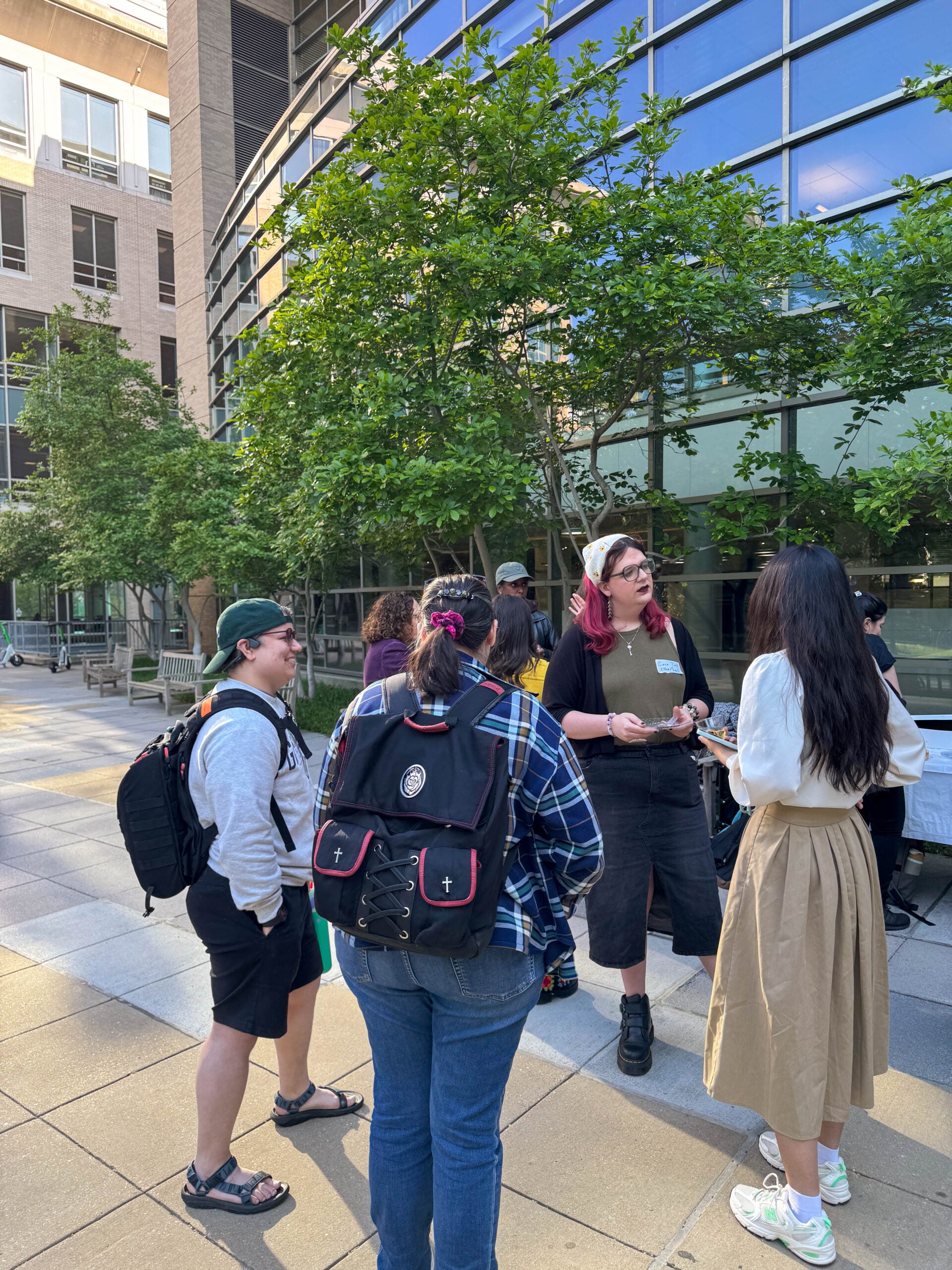 Several people standing outside, talking to each other and holding snacks and cups in their hands. Behind them is a large, tall building with a wall of windows.