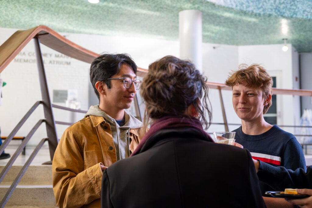  A group of three people standing in a group, holding cups and plates of food. Behind them is a staircase.