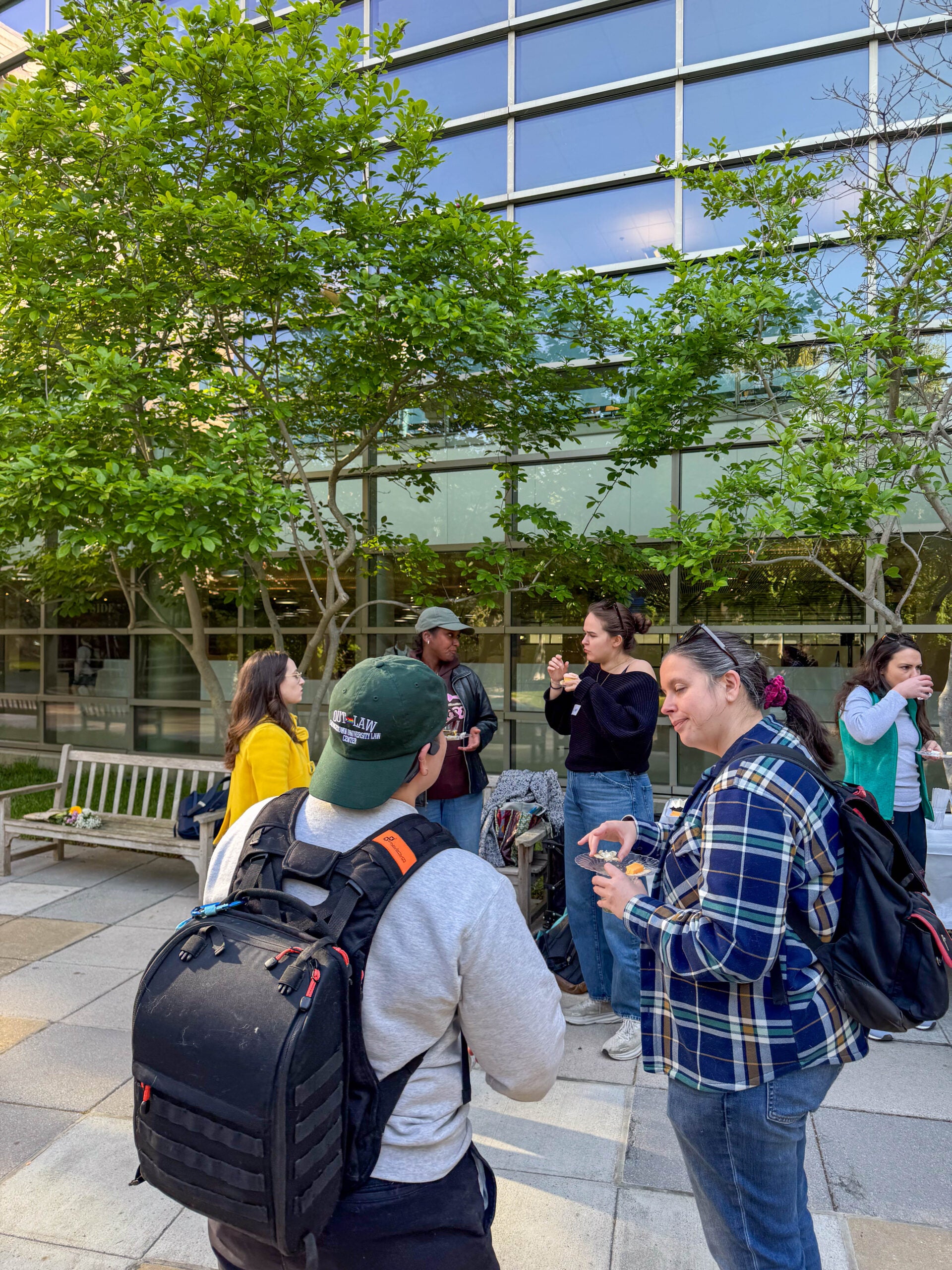 Several people standing outside, talking to each other and holding snacks and cups in their hands. Behind them is a large, tall building with a wall of windows.