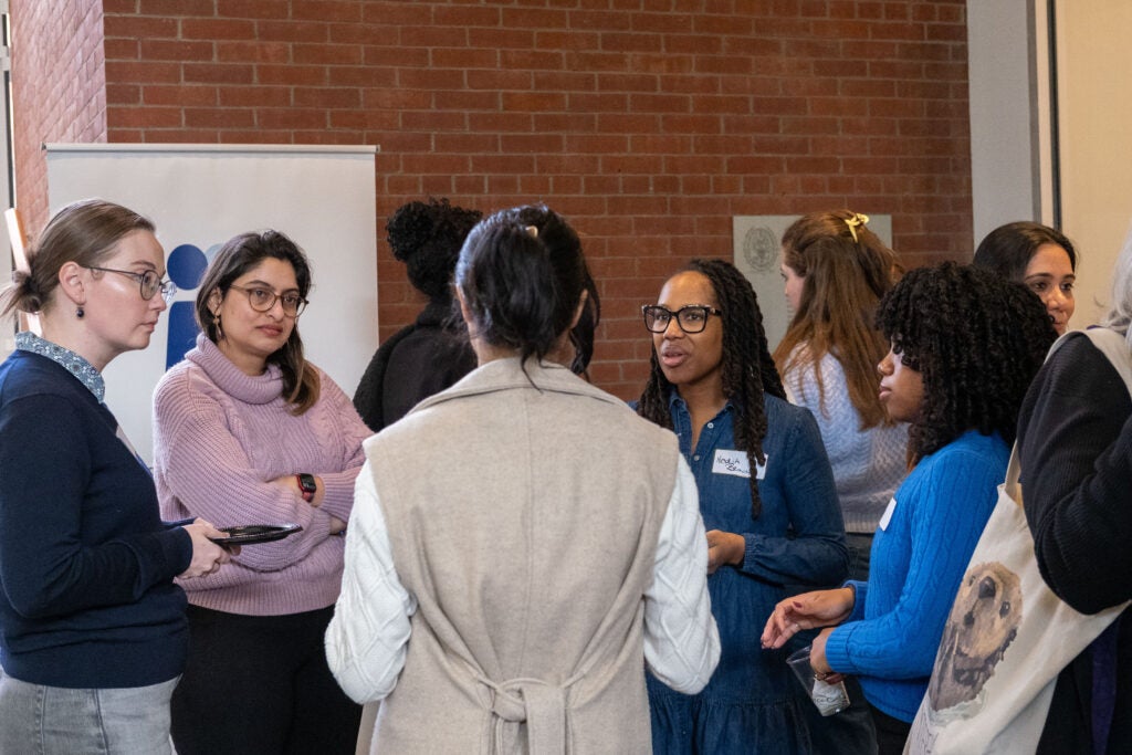 A group of five standing in a circle and talking. Several of them are holding cups and plates of food. In the background is the Gender+ Justice Initiative banner.