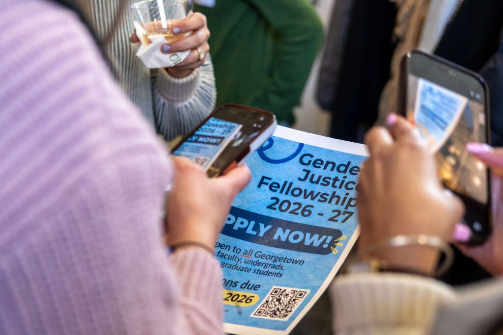 Two people taking photos of a flyer on their cellphones. The flyer is for the Gender+ Justice Fellowship Application.