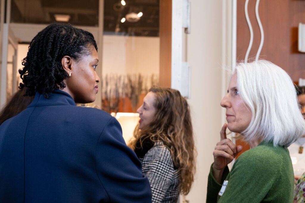 Two women talking to each other. The woman on the left is wearing a blue suit jacket, and the woman on the right holds a finger to her chin and is wearing a green sweater.