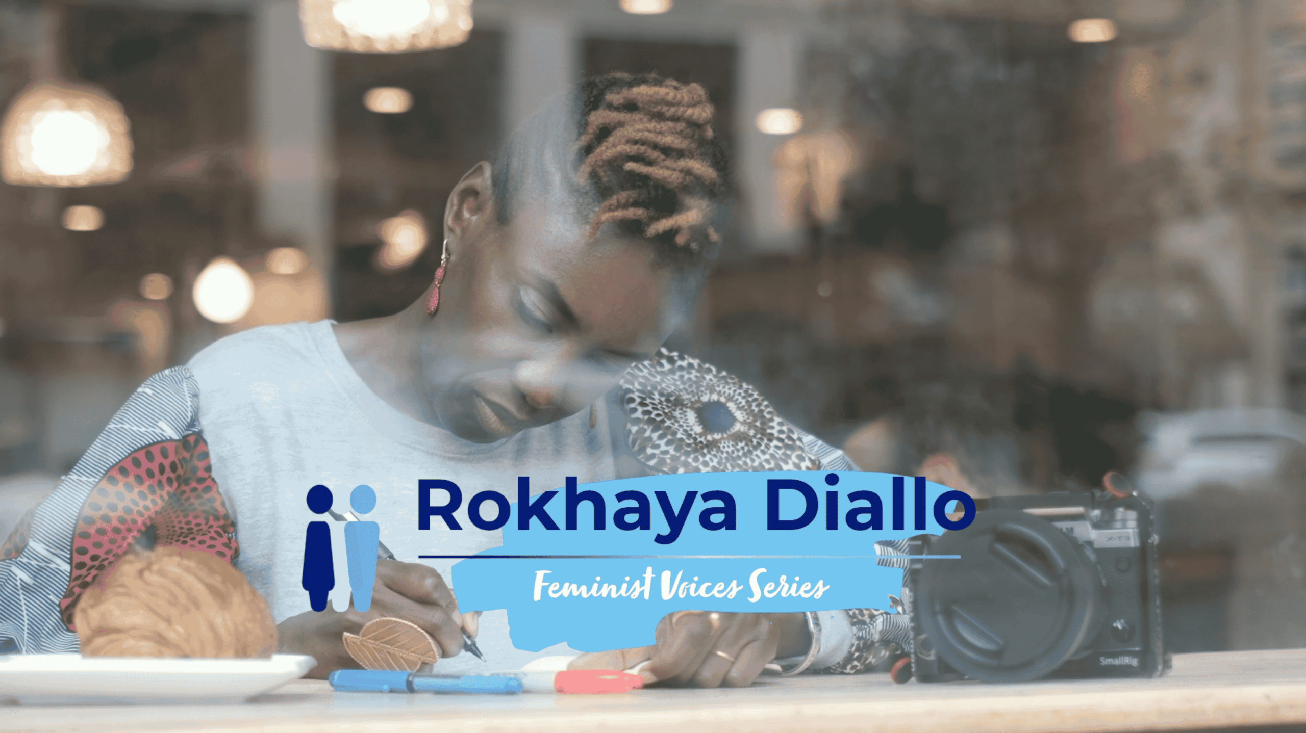 A photo of a woman taken through a window. She is sitting at a table in a cafe, head looking down while writing in a notebook. Blue and white text below her reads 