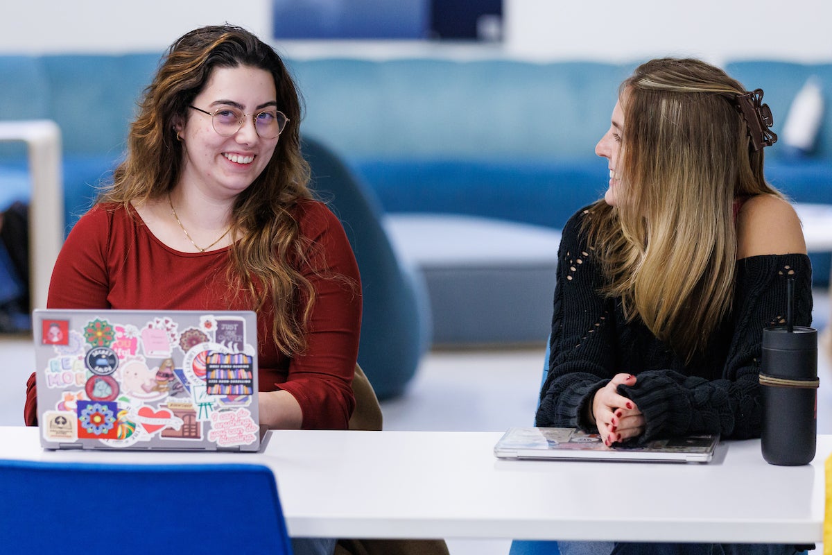 Two women sitting at a table. The woman on the left is smiling and typing on her laptop.