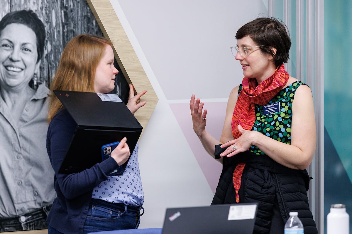 Two women chatting. The woman on the left is holding a laptop computer in her arm.