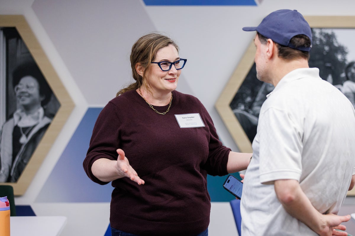 Two people standing and chatting. The woman on the left is smiling and gesturing with her hands. The man on the right has his hands behind his back.
