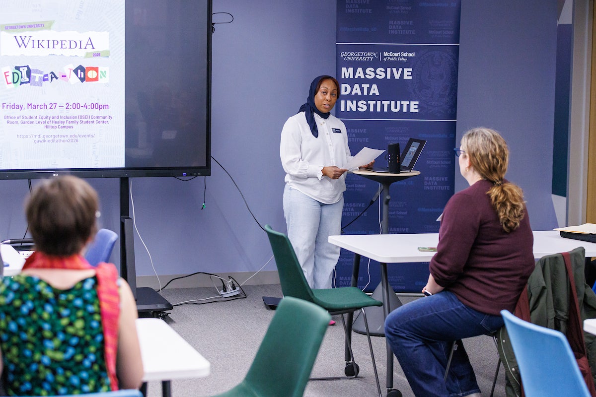 A woman standing at a podium with a sheet of paper, talking to an audience of event participants.
