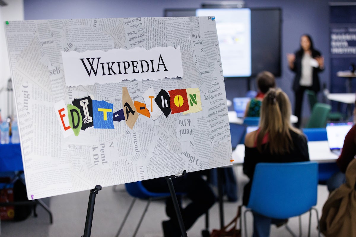 A sign on a pedestal that says Wikipedia Edit-a-thon against newspaper clippings. In the background are event participants sitting at tables.